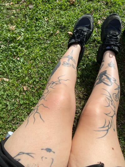 The image shows a person's legs and feet, with black sneakers on. The legs are adorned with numerous black tattoos of various designs, including what appears to be a winged creature and abstract lines. The person is sitting on a grassy area, possibly a lawn or park, with scattered dry leaves and twigs visible in the grass. The lighting suggests it is daytime and sunny. There are no other people or objects of note in the frame. This image does not contain any information that would allow for a specific location in the city of Durazno, Argentina to be identified.