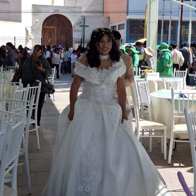 The image captures a wedding celebration in Cerro Colorado, Peru. The bride, adorned in a white gown with floral embellishments and confetti in her hair, is the focal point. Behind her, a crowd has gathered near the entrance of a church with large wooden doors. White chairs are arranged on the street, indicating a reception or celebration. The scene includes spectators, some of whom are wearing green hats, and individuals taking photographs. A banner labeled "Paraguay" is visible, suggesting a connection or reference to the country of Paraguay.