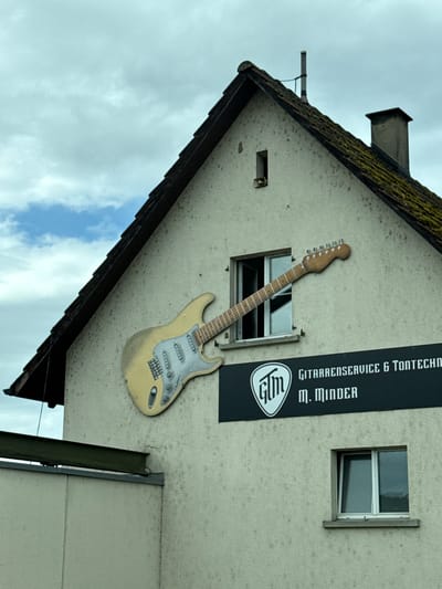 The image captures the exterior of a building in Zurzach, Switzerland, showcasing a unique advertisement against an overcast sky. The building's facade is a light, textured material, possibly stucco, with visible signs of weathering and discoloration.

Dominating the side of the building is a large, three-dimensional decorative guitar, resembling a Fender Stratocaster. The guitar is light yellow or beige with a silver-colored pickguard and pickups, and its neck extends upwards, partially obscuring a small, open window. To the right of the guitar, a tall, black rectangular sign is mounted vertically. The sign features white text, clearly displaying "GITARRENSERVICE & TONTECHNI" (likely 'Tontechnik' for sound technology) followed by "M. MINDER," indicating the name of the business or owner. Below this text, a white logo shaped like a guitar pick with the stylized letters "GLM" or "GTM" is visible.

Further to the right, a smaller, closed window with white frames is set into the wall. The roofline is prominent, featuring dark tiles that appear to be covered in moss or dirt. A chimney stack rises from the roof, and a pipe-like structure is attached to the wall near the left edge of the roof. The sky is uniformly gray and cloudy, suggesting an overcast day. No people are visible, and the scene depicts a commercial building advertising a guitar service and sound technology business.