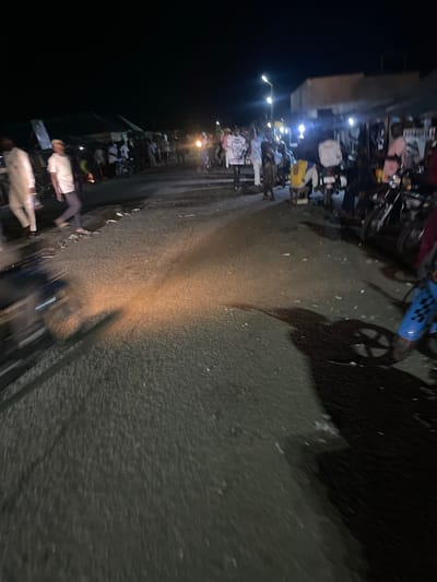 The image depicts a nighttime scene on a paved road in Wai A, Nigeria, likely during a gathering or market event. The road surface appears wet in places, reflecting the artificial lighting from street lamps and stall lights. A crowd of people is visible, with many standing and conversing, while others are on or near motorcycles. Some individuals are wearing traditional Nigerian attire. The background shows stalls and buildings, illuminated by various light sources, creating a lively but somewhat chaotic atmosphere. The foreground shows the road and shadows cast by people and objects. The overall impression is of a busy, active community space at night.