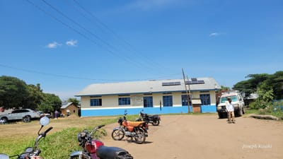 This daytime photograph captures a scene outside a building, identified as a hospital, in Tchere, Democratic Republic of the Congo. The main subject is a light-colored building with a blue stripe along its base and blue-framed windows, situated under a clear blue sky with a few wispy clouds. The roof of the building is corrugated metal and features several solar panels, suggesting off-grid power generation.

A sign on the front of the building is partially legible, displaying "DPS ITURI," "HOPITAL GENERAL DE REFERENCE DE RWAMPAI," and "SERVICES ORGANISES: H."

In the foreground and midground, several motorcycles are parked on a dirt and grassy area. The closest motorcycle, seen partially on the left, has a red tank, while another in the center has an orange seat. To the right of the building, a white vehicle marked "AMBULANCE" is visible, with a person standing near its front. Another white car is parked further left, near a group of people, including children, seated on the ground under a tree.

The ground consists of dirt paths and patches of green grass. Lush green trees are visible on the left and right sides of the frame, indicating a verdant environment. Overhead, power lines stretch across the sky. The lighting suggests it's a sunny day. The watermark "Joseph Kisuki" is present in the bottom right corner.