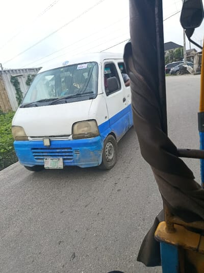 The image depicts an urban street scene in Warri, Nigeria, captured from a moving vehicle, likely a tricycle taxi (keke Napep), indicated by the partial view of a yellow and blue metal frame and dark fabric element in the right foreground.
A white and blue Suzuki Every-style minivan, commonly referred to as a "korope" or "bus," occupies the central midground, moving on an asphalt road. Its top half is white, while the lower half, including the front bumper, is painted blue. The blue paint is faded, chipped, and exhibits dirt and mud splashes. The headlights appear aged. A Nigerian license plate, partially legible and containing "WARRI," is affixed to the front bumper. The driver is visible through the windshield, and multiple passengers are discernible inside, with one person's arm extending from an open rear window on the passenger side.
To the left of the minivan, a weathered corrugated metal fence, partially obscured by green vines and foliage, defines a property boundary. Beyond this, dense green vegetation is visible. To the right, further down the road, a dark-colored vehicle is parked near a stack of tires. Behind these objects, a multi-story building with a dark roof and light-colored walls stands against the horizon. The sky is overcast, and utility lines are visible overhead. The road surface is dry asphalt.