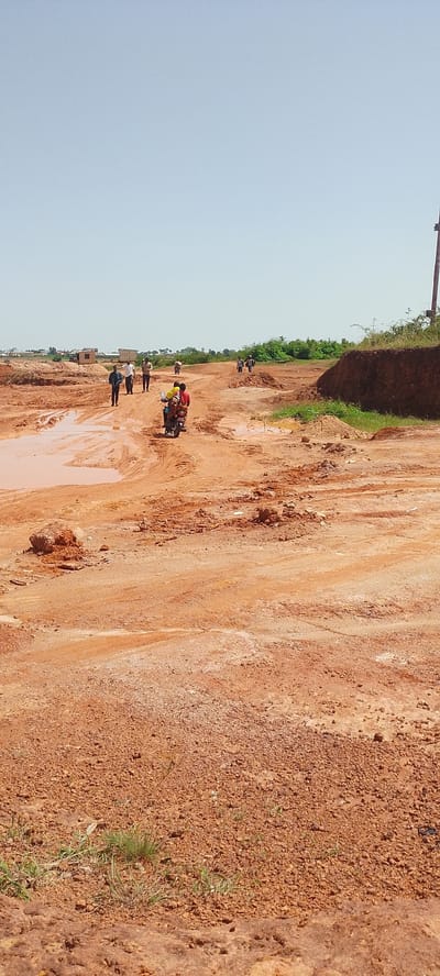 The image depicts a dirt road in Reifild-Ist, Nigeria, characterized by red earth and significant puddles of muddy water, suggesting recent rainfall or poor drainage. Several individuals are present. In the foreground, the terrain is rough and uneven, with loose gravel and clumps of red soil. Further up the road, a group of people are walking in various directions. A motorcycle carrying two individuals, one wearing a yellow shirt and the other a red shirt, is traveling on the road. In the background, more people are walking, and there are scattered structures, possibly buildings, along with green vegetation. The sky is clear and bright blue, indicating a sunny day. The overall scene suggests a rural or developing area undergoing construction or experiencing challenging weather conditions.