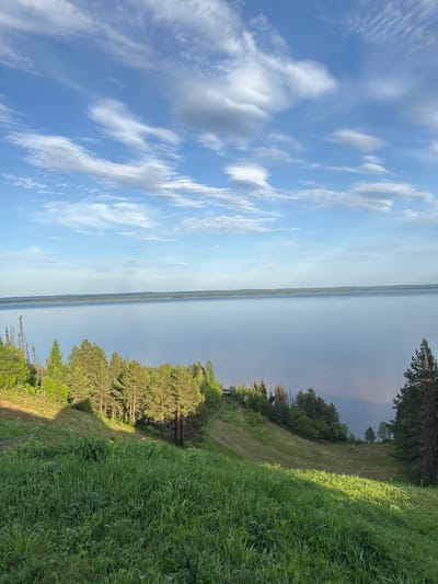 The image shows a landscape in Галево, Russia. The scene is divided into three main sections: sky, water, and land. The sky occupies the left portion of the image, displaying a bright blue color with scattered white clouds. A large body of water spans the middle ground, appearing calm and reflecting the sky's color. On the right, the land is covered with lush green grass and trees, creating a natural setting. The composition appears to be taken from an elevated viewpoint, offering a panoramic perspective of the area. No people or animals are visible in the frame.
