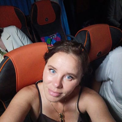 A woman with blue eyes and light skin is taking a selfie on what appears to be a bus. She has brown hair, and is wearing a dark colored tank top and a gold necklace. The woman is smiling faintly as she looks at the camera. Behind her, seats with orange and black upholstery are visible. The lighting is dim, suggesting it is either nighttime or a poorly lit interior. No other individuals are clearly visible, although the arm of another person wearing a white shirt is partially visible on the right side of the frame. The scene appears to be indoors, likely within a vehicle. The location context provided is Svetloe, Russia. There is no visible text in the image.