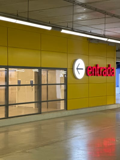 The image shows a downward-pointing arrow within a white, circular sign mounted on a yellow tiled wall. Above the sign, the word "entrada" is displayed in large, red, three-dimensional letters. This signifies an entrance. Below the sign, a large window with a dark frame is visible, revealing the interior of a building. The interior appears to be illuminated, with shelves or displays visible behind the glass. The overall setting is likely a public space, possibly a train station or a public building in Massanassa, Spain, given the context. The lighting suggests it is daytime or that the interior lighting is sufficient. The materials of the wall and the window frame suggest a modern, functional design.
