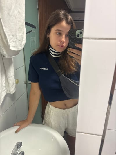 A young woman is taking a selfie in a bathroom. She is wearing a navy blue cropped t-shirt with white stripes at the collar and white speckled shorts. A dark fanny pack is worn across her chest. Her nails are painted a light olive green. She has her left hand resting on the edge of a white sink. A towel is hanging on the left side of the image, and a mirrored door with text that appears to read "HOTEL" is visible in the background. The bathroom tiles are white. The image was taken in Incheon, South Korea.