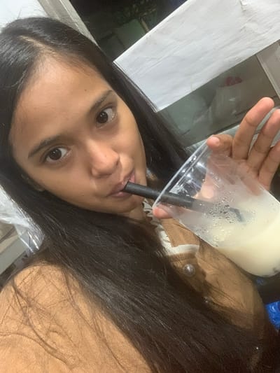 The media file shows a close-up, low-angle shot of a young woman with long, dark hair drinking from a clear plastic cup containing a light-colored beverage. She is holding the cup with her right hand, and a black straw extends from the cup into her mouth. Condensation is visible on the outside of the cup, suggesting the drink is cold. The woman's face is partially obscured by her hair and the cup. Her eyes are open, and she appears to be looking directly at the camera. She is wearing a light brown garment with a textured surface, possibly a sweater or jacket. The background is somewhat blurred, but appears to be indoors or in a covered outdoor area, with a white surface on the left and a dark background on the right. There are no visible signs of specific weather conditions or time of day. The overall impression is that of a casual, personal moment captured. No text is visible in the image.