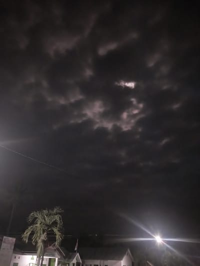 A nighttime outdoor photograph captures a dark, overcast sky filled with heavy clouds. The clouds are dense and appear to be illuminated from behind in some areas, creating a mottled, somewhat ethereal effect. In the lower part of the image, a single palm tree stands prominently on the left, its fronds spread out. Behind and below the palm tree, the roofs and some walls of buildings are visible. One building on the far left has a white facade with some darker rectangular elements, possibly windows. Another building to the right also has a white structure and a sloped roof. A bright light source, likely a streetlamp, is visible on the right side of the frame, emitting a strong glare and lens flare that streaks across the sky. A thin, white line, possibly a wire or a contrail, stretches diagonally across the upper left portion of the image. The overall impression is of a dimly lit, possibly humid, tropical night with thick cloud cover. The scene appears to be set in a residential or semi-urban area, given the presence of buildings and lighting. The location in Labissa, Indonesia, suggests a tropical climate consistent with the palm tree and likely high humidity indicated by the dense clouds.
