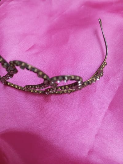 A metallic headband, adorned with small, clear, faceted stones set in multiple linked, decorative segments, rests on a vibrant pink fabric surface. The headband's base appears to be a dull gold or bronze tone. The rightmost end of the thin, curved metallic band extends upwards. No individuals, actions, or specific interactions are visible. The environment consists solely of the pink fabric. Location details for Qarabucaq qəsəbə, Azerbaijan are not observable in the image.