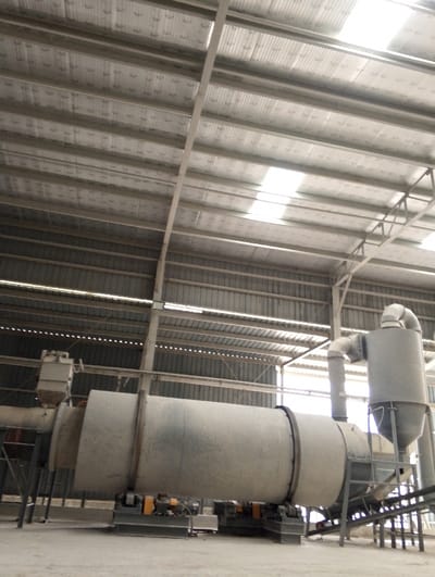 The interior of a large industrial building with corrugated metal walls and a corrugated metal roof, located at coordinates 22.694, 90.601, in Pangasia, Bangladesh. The building appears to be a factory. A large, cylindrical industrial machine, likely a rotary dryer, is situated in the foreground. The machine is supported by a metal frame and appears to have a dust collection system. The machine is surrounded by a concrete floor with evidence of dust. The machine is in the foreground of the image, while the background consists of the building's interior, featuring corrugated metal walls and a corrugated metal roof. There are no people present in the image. The light source appears to be natural, entering through a skylight in the roof. The overall impression of the scene is industrial, with the large machinery and dusty environment suggesting heavy industrial activity.