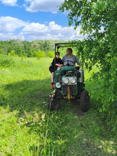 In a grassy field under a bright blue sky with scattered clouds, two individuals are seated on a green tractor. To the right, a mature man with short, graying hair and a grey t-shirt is at the steering wheel. To his left and slightly in front, a young boy wearing a black cap, black hoodie, and black shorts with red and white sneakers is looking towards the camera with a smile. The tractor has two headlights, a yellow tow hitch, and large black tires. The background is filled with lush green trees and vegetation. The location is identified as Ilmino, Russia.