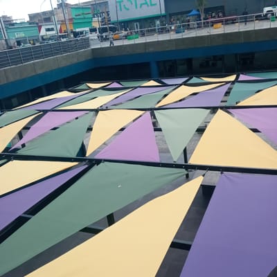 The image shows a colorful canopy of triangular fabric shades in shades of yellow, purple, and green, suspended over an outdoor area. These shades are attached to a dark metal framework. Below the canopy, a grey concrete structure with blue accents is visible, and further up, a street scene unfolds. In the background, buildings with storefronts are present, including one with a large "TOTAL" sign. People are seen walking on the street, and vehicles, including cars and a white truck, are also present. The overall setting appears to be an urban environment, possibly a commercial area or a public space in El Limon, Venezuela. The lighting suggests it is daytime.