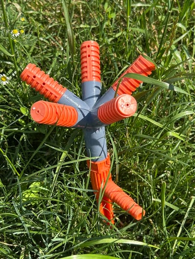 The image shows an orange and grey garden sprinkler lying on the ground amidst lush green grass and a few small white wildflowers. The sprinkler is star-shaped, with multiple arms extending outwards. Each arm is orange and ribbed, with small circular openings near the tip. The central hub of the sprinkler is grey. The grass is tall and dense, with some blades casting shadows, suggesting it is a sunny day. The overall setting appears to be a garden or a grassy field in a rural location. There is no text visible in the image. The scene is peaceful and appears to be outdoors during daylight hours. The sprinkler is not actively in use; it is lying on its side. There are no people or animals present in the frame. The focus is on the garden sprinkler and its natural surroundings. The image evokes a sense of summer or late spring due to the vibrant green grass and blooming flowers. The lighting suggests it could be late morning or early afternoon.