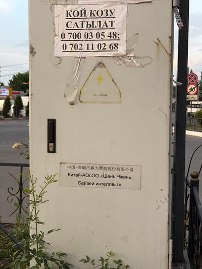 This is a photograph of a metal enclosure with several stickers and labels. The enclosure appears to be outdoors, with a fence and some greenery visible in the background. There are two prominent stickers on the left side of the enclosure. One has text in Cyrillic and appears to be advertising something with phone numbers: "КОЙ КОЗУ САТЫЛАТ 0 700 03 05 48; 0 702 11 02 68". Another sticker is mostly obscured but contains some red and black text.
On the right side of the enclosure, there is a white label with Chinese and Russian text. The Chinese text reads "中国-深圳市赛为智能股份有限公司", which translates to "China-Shenzhen Sifang Smart Co., Ltd.". Below this, the Russian text states "Китай-АОСОО «Шень Чжень Сайвей интеллект»", which appears to be a transliteration and translation of the company name.
There is also a yellow sticker with a black lightning bolt symbol, indicating danger or electricity. Below this symbol, in Chinese characters, it says "止步:有电危险", meaning "Stop: danger of electricity". A black lock mechanism is also visible on the enclosure.
In the background, there are traffic signs, including a circular sign with a red border and a diagonal red bar, and another sign with the number "40" inside a circle. The overall scene suggests an industrial or utility area, possibly near a road. The lighting suggests it is daytime. The presence of multiple languages on the labels indicates an international context or a place with diverse linguistic influences.