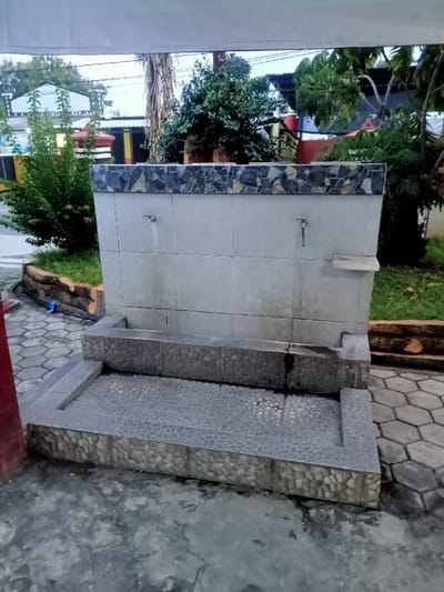 The image shows a small, grey concrete wash station with two faucets, located in the Kecamatan Margorejo district of Surabaya, Indonesia (latitude: -6.737, longitude: 111.014). The wash station has a small trough at the bottom, which appears to be wet, suggesting recent use. The wash station is made of several sections of grey, textured tiles. The faucets are in a slightly recessed alcove above the trough. There is a small shelf on the wall to the right of the right faucet. The wash station is surrounded by a paved area, which appears to be grey concrete with hexagonal pavers. The background includes lush green foliage, and a portion of a white, canvas awning can be seen in the upper left corner of the image. There are no people visible in the image.