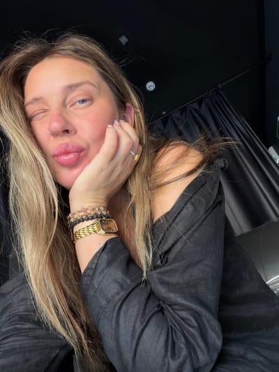 The image shows a close-up of a woman's face, tilted to the left, with her right eye closed in a wink and her left eye open, looking forward. Her lips are pursed, and her right hand is resting on her cheek, with her thumb supporting her chin. She is wearing several bracelets on her left wrist, including beaded bracelets and a gold watch with a square face. A gold ring with a yellow stone is visible on her right ring finger. Her hair, which is blonde and reaches past her shoulders, is partially obscuring her face and body. She is wearing a dark-colored garment, possibly a shirt or dress, with a textured fabric. The background is dark and appears to be a draped curtain or fabric. There is no discernible city or location information visible in the image.