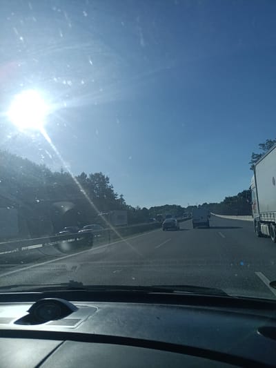 The image captures a view from inside a vehicle on a highway, likely in Kunice, Czechia, on a sunny day. The sun is bright and positioned in the upper left quadrant of the image, creating a lens flare and casting strong light across the scene. The sky is clear and blue, suggesting fair weather.
The highway appears to have multiple lanes, and traffic is visible. Several cars and vans are on the road, some moving, others appearing to be in slow-moving traffic. A large white truck is prominent on the right side of the image, its trailer bearing a logo with a red circular element and possibly text in English, as indicated by the Union Jack flag design on the side. On the left side of the highway, there is a steep embankment covered in trees and vegetation, with a concrete barrier separating the road from the embankment. Some structures are visible on the embankment, potentially part of an overpass or noise barrier.
The dashboard of the vehicle is partially visible in the foreground, showing a circular gauge or speaker. The windshield has some streaks or smudges, further indicating it's a view from inside a car. The overall impression is of a typical highway journey on a bright, clear day, with normal traffic conditions. The time of day is likely late morning or early afternoon, given the angle and intensity of the sun.