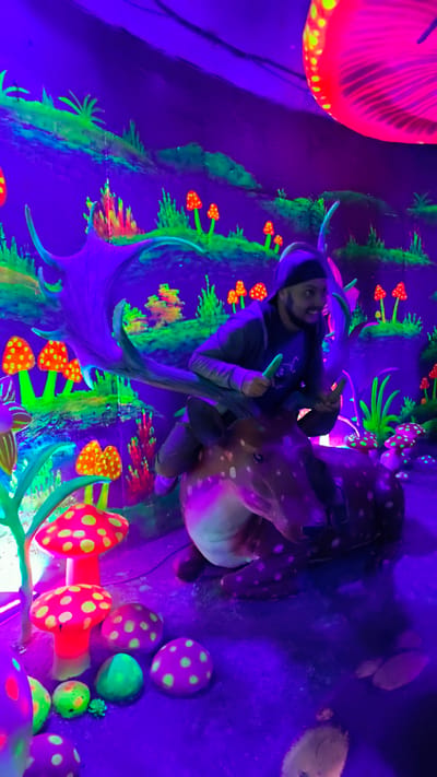 A person is seated on a deer sculpture within a black light exhibit. The person wears a dark beanie and a grey jacket and holds two green objects. The environment is a whimsical forest landscape with painted elements including fungi, plants, and mushrooms. The colors in the image are predominantly violet, pink, and green, with a blacklight effect making the colors vivid. The photo was taken in Binh Lu, Vietnam.