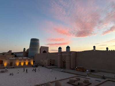 The image shows a panoramic view of Khiva, Uzbekistan, during sunset. The sky is filled with vibrant pink and orange clouds, indicative of the setting sun. In the foreground, a large courtyard is paved with stone. Several people are scattered across the courtyard, with some children playing and adults walking. Two bicycles are visible, one of which has a rider. To the left, a building with illuminated windows is prominent. In the background, a massive, intricately decorated blue minaret stands tall, alongside other traditional buildings and walls characteristic of Khiva's historic architecture. The overall scene depicts a lively evening in the ancient city.