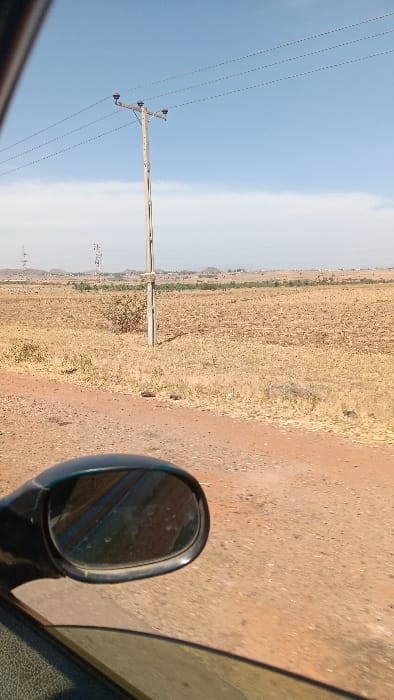 The image depicts a dry, open landscape in Barakin Tenti, Nigeria, viewed from inside a vehicle. The foreground features a reddish-brown dirt road, with the black-cased side mirror and a portion of the window frame of the vehicle partially obscuring the lower left. The environment beyond the road consists of vast, dry, yellowish-brown fields with sparse, small, dry bushes. In the mid-ground, a tall, light-colored utility pole with a crossarm supports three power lines, which extend horizontally across the upper portion of the frame. Further in the mid-ground, to the left of the pole, two distinct metal lattice communication towers are visible. The horizon shows distant, hazy hills or mountains. The sky is a light blue with scattered white clouds, indicating daylight conditions. No people are visible.