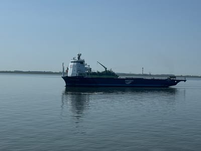 A large, dark blue barge named "EASTERN HUSK" is moving across calm, rippling water under a clear, bright sky, suggesting daytime with fair weather. The barge has a prominent white superstructure at the front, housing what appears to be a bridge or control room, complete with radar dishes and antennas. A green crane is visible on the deck of the barge. The word "HARTMAN" is emblazoned in white on the side of the barge, indicating the company operating it. In the distance, a thin line of land with trees and structures is visible, hinting at a coastline or riverbank. There are also some tall, industrial-looking structures in the far distance, possibly lighthouses or other navigational aids. The water is calm and reflects the sky and the barge, creating a serene and peaceful atmosphere. The overall scene depicts maritime activity in a coastal or estuarine environment. Given the location context of Brunsbüttel, Germany, the scene likely takes place on the Elbe River or the Kiel Canal.