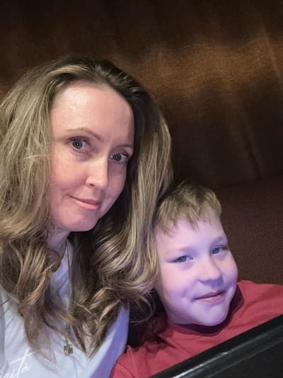 A selfie-style image shows a woman and a young child lying down. The woman has long blonde hair, light skin, and blue eyes. The child also has light skin, blonde hair, and blue eyes and wears a red shirt. The background is a dark brown surface. There is no indication of a location in Balezino, Russia.