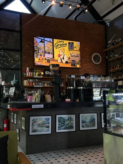 The image depicts the interior of a cozy cafe, likely in Prachuap Khiri Khan, Thailand, as suggested by the context. The scene is captured from a slightly elevated, tilted perspective, giving a dynamic feel to the shot. The cafe features a rustic brick wall as a prominent backdrop, adorned with a decorative circular mirror.

A large, vibrant sign with "CAFE Groove" written prominently is displayed on the brick wall, featuring stylized illustrations of coffee making and a joyful figure. Below the sign, on a counter, are coffee grinders and various coffee-related products. Behind the counter, a person's head and shoulders are visible, suggesting they might be a barista or staff member.

The counter itself is cluttered with various items, including coffee cups, packets, and what appear to be menu displays. Two framed watercolor paintings of natural landscapes hang on the dark wall behind the counter, adding an artistic touch. The cafe also has modern lighting fixtures and large windows that allow natural light to enter, though the time of day isn't explicitly clear.

Visible text includes a menu with coffee prices and types such as "DRIP COFFEE," "AMERICANO," "ESPRESSO," "CAPPUCCINO," and "CAFE LATTE." Other text includes "CAFE GROOVE," "Chill Jazzhop & Groove Pop," "GOOD COFFEE," "GOOD MUSIC," and "GOOD DAY." Additionally, there's a sign in Thai that reads "สั่งอาหารตรงนี้," which translates to "Order food here." The overall atmosphere appears relaxed and inviting, indicative of a pleasant cafe experience.