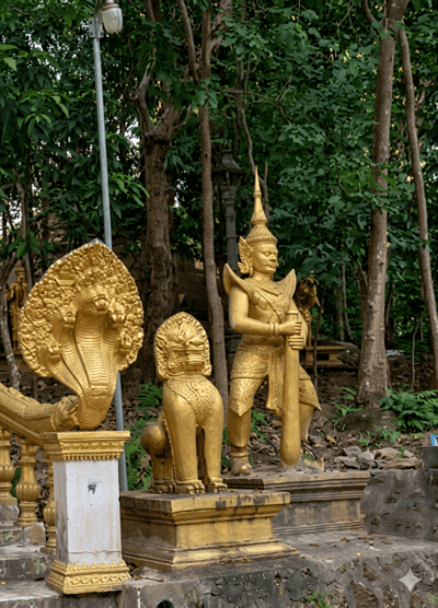 In Sihanoukville, Cambodia, amidst a lush forest setting, a series of golden statues are displayed outdoors. The main subjects are three distinct golden sculptures. To the left, a prominent statue depicts a multi-headed Naga, a mythical serpent, with detailed scales and ornate decoration. Adjacent to it, to the right, stands a golden statue of a mythical lion-like creature, a Chinthe, with a fierce expression and ornate detailing. Further to the right and slightly behind, a larger golden statue of a warrior figure is presented. This figure is adorned with a tall headdress and holds a long, cylindrical object that appears to be a weapon. The statues are placed on golden pedestals, with the Naga and Chinthe statues appearing to guard a walkway or entrance, indicated by the presence of a railing to the left. The environment is characterized by tall trees with dense green foliage, and the ground is covered with leaves and scattered stones. A street lamp with a white fixture is visible on a pole to the left, suggesting an outdoor, possibly public, location.