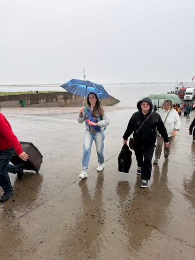 The image depicts an outdoor scene on a rainy or recently rained-on day, with a uniformly grey, overcast sky. The ground is a wet, paved surface, possibly a dock or pier, showing reflections of the people and the environment.

In the midground, a young woman stands holding a blue umbrella adorned with white star-like patterns. She is wearing a denim jacket, light blue jeans, and white sneakers, with a "NIKE" fanny pack across her chest, and she has a slight smile. Next to her, a man in a black hooded sweatshirt, dark pants, and sunglasses walks forward, carrying a black bag in one hand and a shoulder bag. Behind him, an older woman in a white puffer jacket and light-colored pants holds a light green umbrella. In the bottom-right corner, a person wearing a red top and dark jeans is partially visible, holding a black suitcase.

In the background, beyond the paved area, a wide body of water stretches into the distance, with a faint landmass visible on the far horizon. On the right side of the background, several trucks and what appears to be part of a large vessel or structure are visible, along with other people. The overall atmosphere is cool and damp.