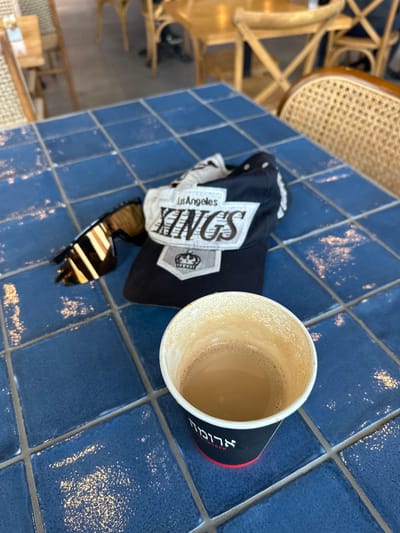 The image captures a scene at Aroma Espresso Bar in Gan Yavne, Israel. On a blue tiled table sits a nearly empty cup of coffee, a black Los Angeles Kings baseball cap, and a pair of sunglasses. The table is positioned near wicker chairs, and the background reveals a partial view of the restaurant's interior. The lighting suggests it's daytime.