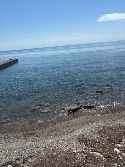 The image depicts a shoreline in Semidvorye, Ukraine. The scene features a beach covered in pebbles and small rocks transitioning into the blue waters of the Black Sea. The sea extends into the horizon under a clear blue sky with a solitary white cloud. A concrete structure, potentially a pier or breakwater, is visible extending into the water on the left side of the frame. The water is clear near the shore, allowing visibility of rocks beneath the surface. No people are visible in the image.