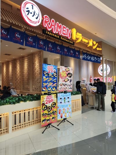 The image shows the interior of a Japanese restaurant called "Ramen 1" in Telukjambe, Indonesia. The restaurant's exterior is visible in the background, with its name and signage clearly displayed. There are several patrons sitting at tables. In the foreground, three menu boards stand on black tripods with their prices and descriptions prominently displayed. Two staff members are present, one in the foreground, and the other in the background, both are wearing red t-shirts. They are interacting with customers. The restaurant's interior is decorated with wooden walls and features a patterned design. The floor is tiled, and the lighting is bright. The overall atmosphere appears clean and inviting.  The restaurant is situated in a mall, with the entrance to the mall visible in the background.  The coordinates are consistent with the location.