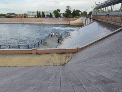 The image depicts a scenic view of the Tobol River embankment in Kurgan, Russia. A concrete embankment, paved with grey granite slabs, slopes down towards the river. The river flows with visible currents and white water. On the left side of the image, a walkway paved with yellow square tiles is visible, separated from the river by a black metal railing. A group of five people are standing near the railing, some appearing to be fishing. Across the river, a long, multi-story building with a green roof and a blue facade stands against a cloudy sky. Further in the background, trees and other smaller buildings are visible. A bridge with lampposts and cars is seen spanning the river in the upper right portion of the image.
