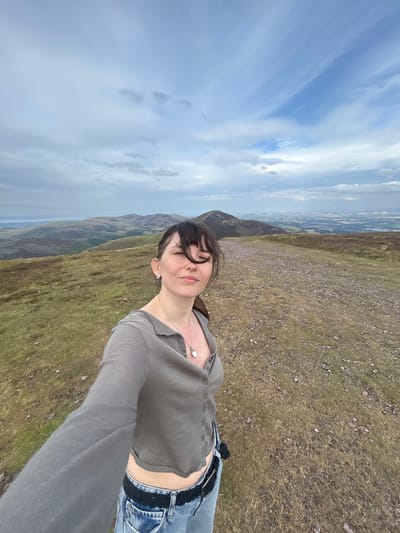 A woman is taking a selfie on a grassy hillside overlooking a valley with a town in the distance, likely near Penicuik, United Kingdom. The woman, with dark hair and bangs, wears a gray cropped shirt with buttons and blue jeans. She has a necklace with a white pendant and earrings. The landscape consists of rolling hills covered in dry grass and heather, with a dirt path leading away from the viewer. The sky is cloudy with patches of blue. The overall scene suggests an outdoor recreational activity in a rural setting close to an urban area.