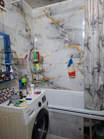 This is a medium shot of a cluttered bathroom in Ridder, Kazakhstan. The walls are tiled with a marble-like pattern featuring gold veining. A white bathtub is partially visible, with a grey and white marble-patterned shower curtain drawn to the right. To the left, a washing machine is positioned next to the bathtub. On top of the washing machine, there are various toiletries and cleaning supplies, including spray bottles, containers, and what appears to be a pack of cigarettes. A shelf unit is mounted on the tiled wall above the washing machine, holding more bottles and containers. A showerhead and faucet are visible near the shelf unit. A towel with red, white, and blue stripes hangs from a towel rack on the wall. The overall impression is of a functional but somewhat disorganized bathroom space.