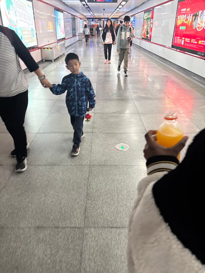 A child walks in a subway holding a rose. An adult accompanies the child by holding their hand. In the foreground, a hand holds a bottle of juice. Several other people are walking in the background, and advertisements are seen on the subway walls. The floor is gray tile, and the lighting is fluorescent.
