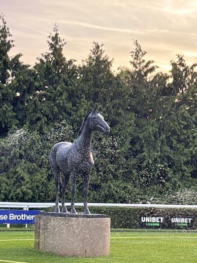 A sculpture of a horse stands on a block of stone, with water spraying upwards around it. The statue and fountain are set against a backdrop of trees and a cloudy sky. In the foreground, there is a sign with the words "Unibet" and "BetMGM", as well as "Chase Brother" on the right side of the image, which could suggest this is at a racetrack or equine event. The location is the Borough of Spelthorne, United Kingdom.