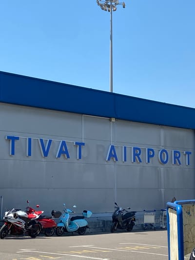 The image depicts the exterior of an airport terminal building, with large, blue and white letters spelling out "TIVAT AIRPORT" vertically on the grey facade. The building features a prominent dark blue overhang or structural element. In front of the building, parked along a paved area marked with yellow lines, are several scooters and motorcycles. There is a light blue scooter, a red scooter, and a white and red motorcycle. A dark grey scooter is also visible. Two luggage carts are parked near a blue railing in the upper right portion of the image. The sky is clear and blue, suggesting it is daytime and the weather is fair. The scene appears to be an airport entrance or waiting area, possibly for departures or arrivals. No people are visible in the image. The location context is Mrčevac, Montenegro.
