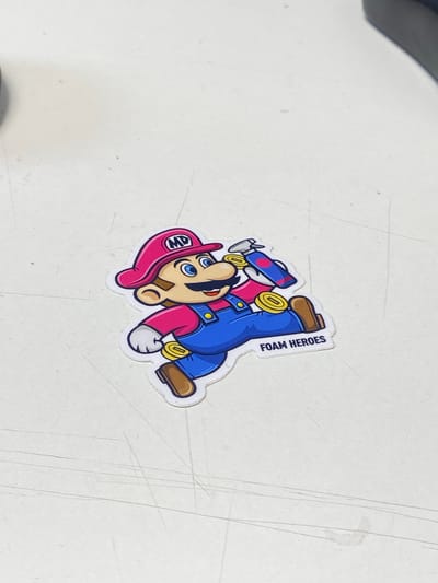 A sticker depicting a cartoon character resembling Mario, wearing a pink hat with "MD" on it, a pink shirt, and blue overalls, is placed on a white surface. The character is holding a blue spray bottle with pink accents. The sticker has the text "FOAM HEROES" printed on it. The white surface has faint lines and specks on it. No information about the city of Uchaly, Russia, is present in the image.