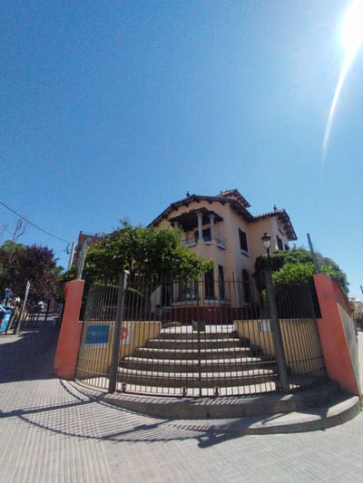 This photograph depicts a grand, light-colored building in Rubí, Spain, under a clear and intensely bright blue sky. The multi-story house, possibly a historic villa, features a tiled roof, decorative eaves, dark window shutters, and a prominent balcony supported by columns. It is situated on an elevated plot, accessed by a wide, curving set of concrete steps leading up from the street level. A tall, dark metal fence encloses the property, flanked by reddish-orange low walls. Lush green trees and bushes are visible around and behind the house.
The scene is bathed in strong sunlight, with a prominent lens flare in the upper right corner of the frame. The bright sun casts distinct, long shadows of the fence onto the patterned, light-colored tiled sidewalk in the foreground, indicating a daytime setting, likely mid-morning or early afternoon. No people are visible. A street lamp post stands near the right side of the fence. A partially obscured blue sign behind the fence appears to contain the text "24H". Faint characters resembling "B1" or "31" are visible on the reddish-orange wall on the right. In the background to the left, additional trees and power lines suggest a residential street.