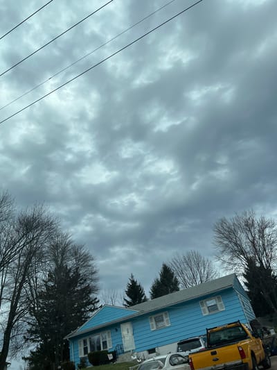 The image depicts a residential scene in the Town of New Windsor, United States of America, under an overcast sky filled with thick, grey clouds. Utility lines traverse the upper portion of the frame. A light blue single-story house with a grey roof, white trim, and a white front door is centrally positioned. White shutters are present on multiple windows, and a window-mounted air conditioning unit is visible on the left side of the house. A short black-railed stairway leads to the front door. Two white lawn ornaments, resembling geese, are situated on the green lawn near the steps, alongside an illegible sign. Bare deciduous trees are prominent on both the left and right, with denser evergreen trees visible further behind the house. Three vehicles are parked in front of the residence: a yellow pickup truck equipped with a bed rack on the right, a white sedan to its left, and a partially visible white vehicle (likely an SUV or van) behind the sedan and pickup. No individuals, explicit actions, or interactions are visible.