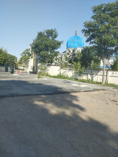 The image shows a mosque with a blue dome in the city of Kyzyltog (Krasnogorsk), Uzbekistan. The mosque is surrounded by trees and a white wall. In the foreground, there is a paved area with some gravel. The sky is clear and blue. The scene appears to be calm and quiet. There are no people visible in the image.