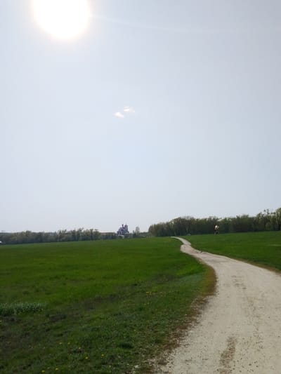 The image depicts a rural scene in Khlevnoye, Russia. A dirt path curves through a grassy field, leading towards a distant building with a prominent red roof, likely a church. The sky is bright, with a visible sun and a small cloud. A line of trees borders the edge of the field. A bird sits on the path.