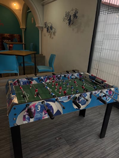 The image shows a foosball table in a room with a quirky decor. The foosball table is blue with images of soccer players and soccer balls printed on its sides. It has the typical rows of foosball players in red and blue. The table is placed on a wood-like floor that is grey. Overhead, there's a ceiling with marine-themed decorations. In the background, one can see an archway leading to an area with blue chairs and patterned cushions. The image was taken inside the Container City complex in San Pedro Cholula, Mexico.