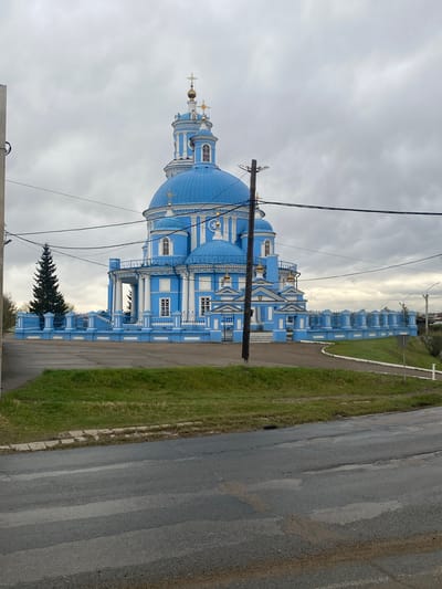 The image depicts the Church of the Kazan Icon of the Mother of God in Telma, Russia. The church is primarily blue and white with multiple domes. It is situated near a road and a grassy area. Power lines are visible in the foreground. The sky is overcast.