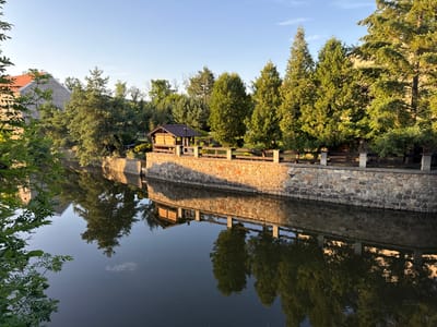The image depicts a serene waterside scene bathed in the golden light of late afternoon or early morning. A stone retaining wall lines the edge of a calm body of water, its rough-hewn stones casting a warm glow. Above the wall, a wooden fence separates the embankment from a line of lush green trees and vegetation. A small, rustic wooden structure with a brown roof, possibly a gazebo or shed, is situated behind the fence. Tall, coniferous trees dominate the upper part of the scene, their vibrant green foliage contrasting with the clear, pale blue sky. The water reflects the trees, the wall, and the sky, creating a mirror image that softens the overall composition. A portion of a building with a red tile roof is visible on the far left, peeking through dense green leaves. There are no people or animals visible in the frame. The scene evokes a sense of tranquility and natural beauty. This location is likely part of the Berounka River or a park area within the city of Beroun, Czechia, given its picturesque riverside setting and well-maintained landscaping.