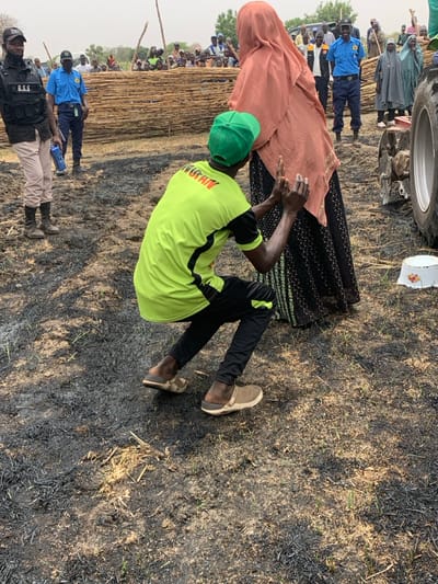 A man wearing a green shirt with the word "AGITHN" printed on it is helping a woman wearing a pink hijab get up off the ground. The scene appears to take place in an open field with some burned grass, near a tall wooden fence and a parked tractor. The location is Ringimawa, Nigeria at 11.666° N latitude and 8.416° E longitude. The man and the woman are the only people in focus.
