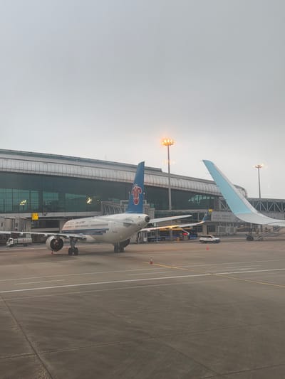 The image shows a China Southern Airlines aircraft parked at a gate at Guangzhou Baiyun International Airport in Baiyun District, Guangzhou, China. The aircraft is positioned adjacent to a terminal building with glass windows. Ground crew vehicles and equipment are visible near the plane. The sky is overcast.