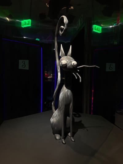 The photograph captures a dimly lit interior, possibly a museum or an exhibit. In the foreground, a life-size, stylized sculpture of a cat is centrally placed on a dark, flat surface. The cat sculpture is monochromatic, with a textured, almost metallic gray finish. It is depicted in a seated position, with large, wide-set eyes, pointed ears, and long whiskers. Its tail curves dramatically upwards, forming a large loop. The sculpture's design is somewhat unsettling, with exaggerated features.
Behind the sculpture, the environment is dark, with hints of colored lighting. On the left and right walls, there are dark doorways with vertical blue and red light strips respectively, creating a dramatic ambiance. Above these doorways, illuminated green exit signs are visible, featuring the standard running-man pictogram. On the dark walls, subtle numbers are displayed; a "3" is visible on the left wall, and a reversed "E" or "3" is on the right wall.
Reflections are present on the ceiling and possibly on the walls, indicating a glossy or mirrored surface. In the background, through the doorways, other figures and elements are dimly discernible, including a person wearing a red shirt, suggesting other visitors or staff. The overall scene evokes a sense of theatricality and artistic presentation, with the lighting and the unique sculpture as the primary focal points.
This image was taken in the Museo de Arte Contemporáneo de Monterrey (MARCO), located in Monterrey, Mexico.